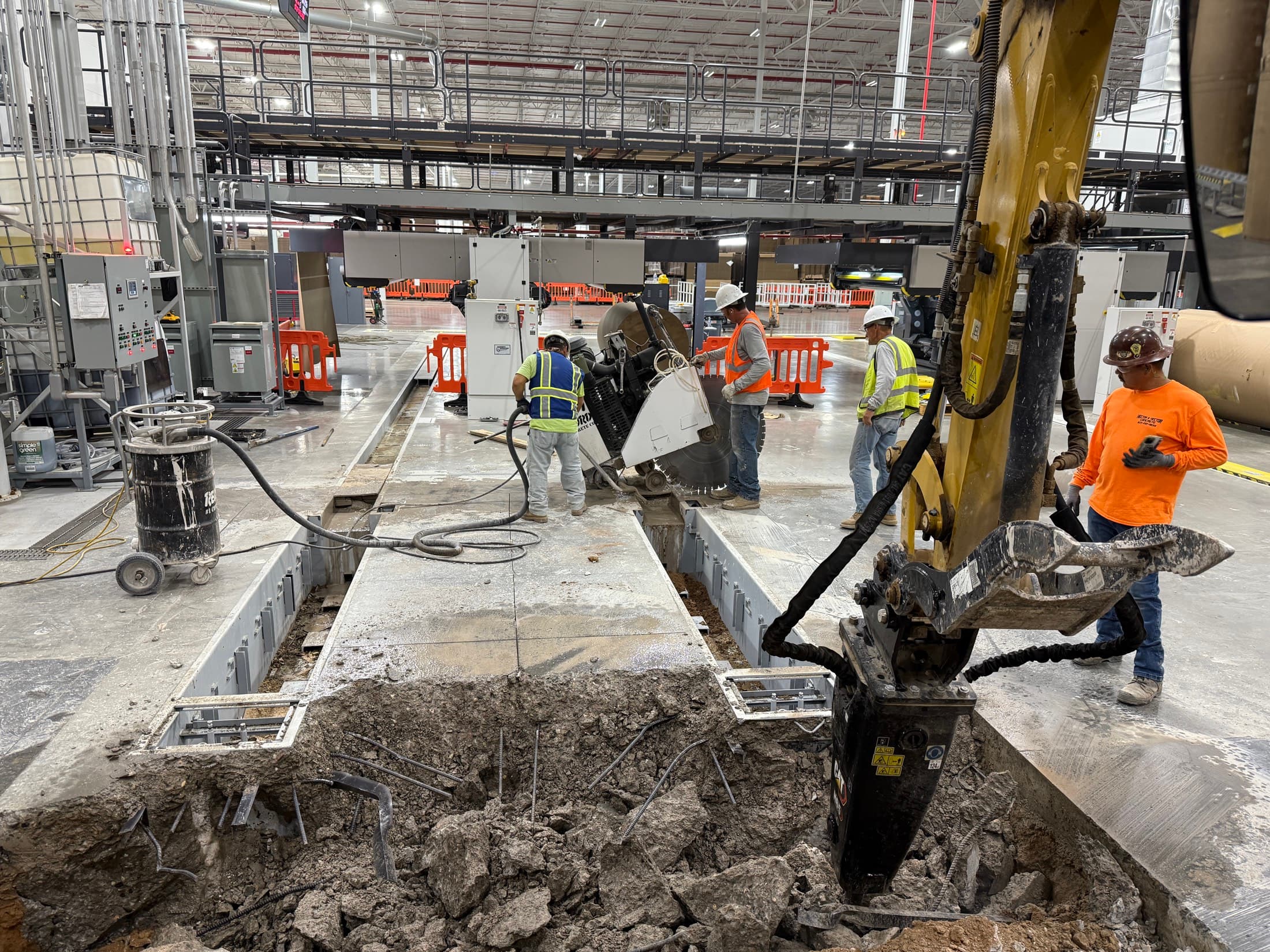 Excavator-mounted hammer breaking out floor beside installed trench steel inside a high-bay plant