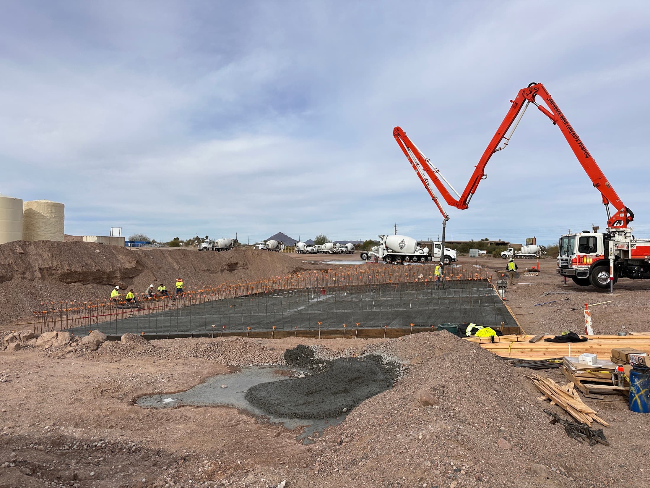 Two concrete pump booms set up over a formed foundation with a line of mixer trucks waiting